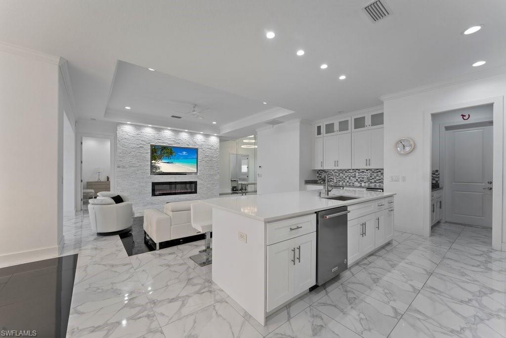 Open-concept living space featuring large format porcelain tile flooring, a kitchen island with a built-in stainless steel dishwasher, white cabinetry with upper glass-front cabinets, and a mosaic tile backsplash