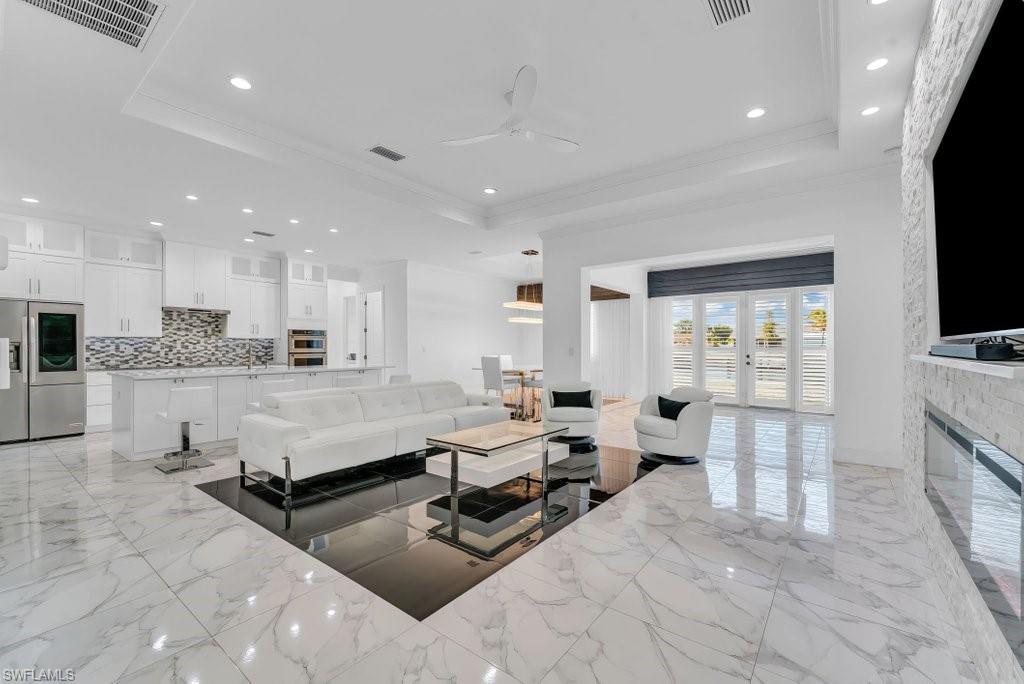Open-concept living space featuring high ceilings with recessed lighting, an integrated kitchen with stainless steel appliances and white cabinetry, a built-in fireplace with a stone surround, and large French doors with interior shutters