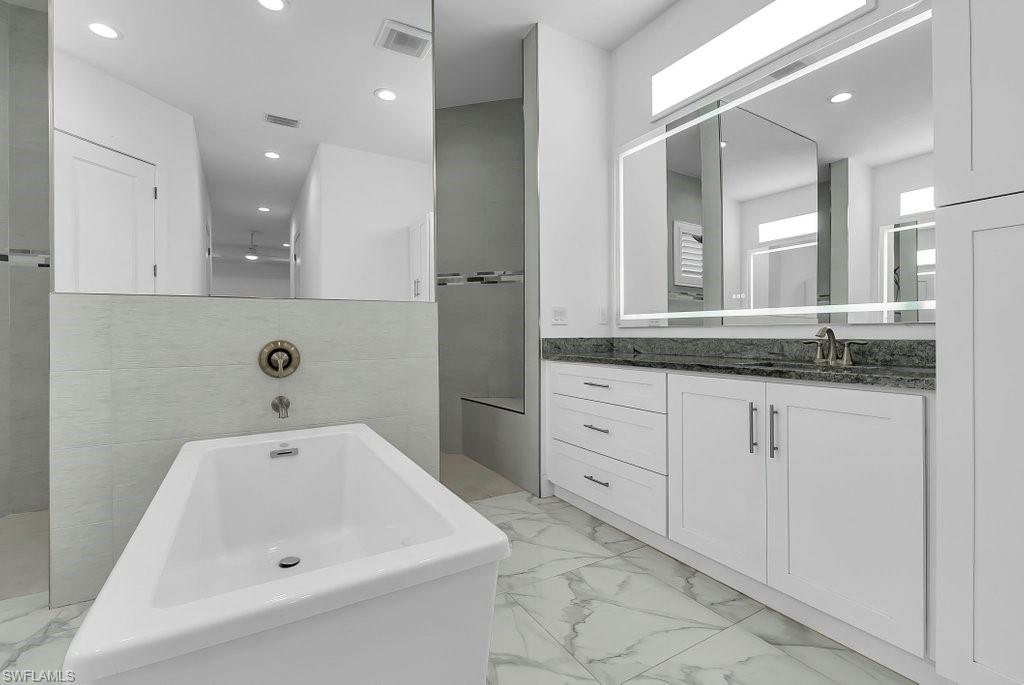 Contemporary bathroom featuring a freestanding soaking tub, large vanity with dark countertop, integrated lighted mirror, and marble-look floor tiling