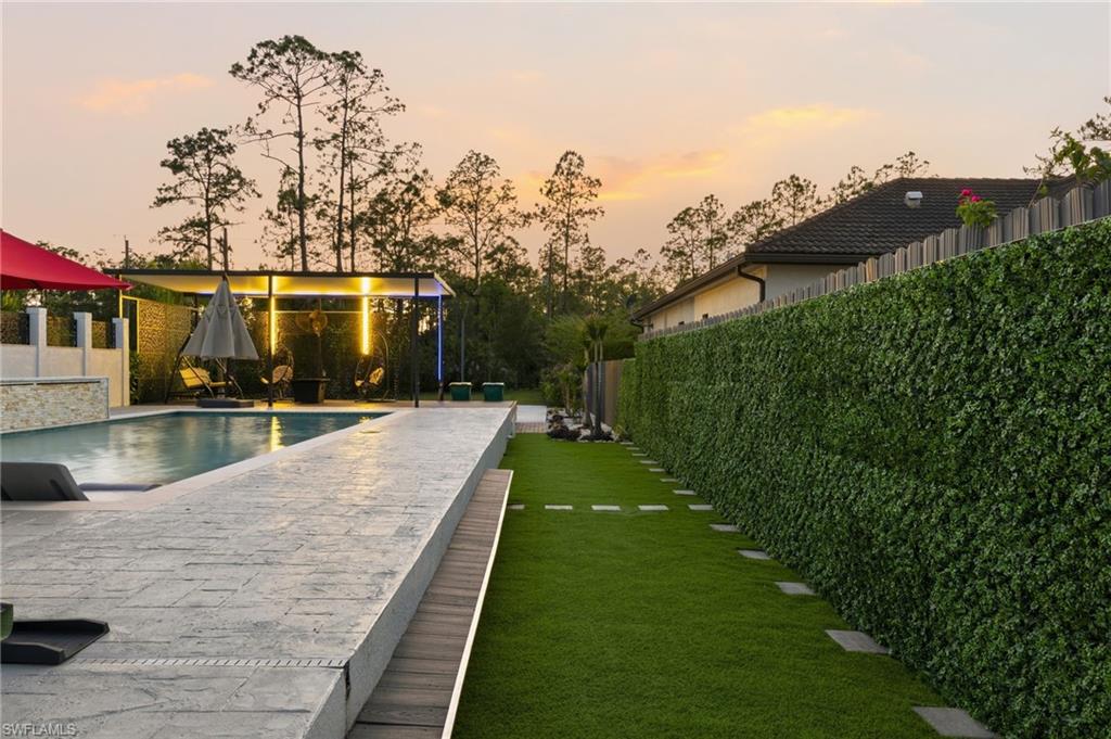 Expansive backyard featuring a saltwater swimming pool with waterfall, a covered patio area with built-in lighting, a stone-tiled pool deck, a lush green lawn with stepping stones, and a tall, verdant privacy hedge