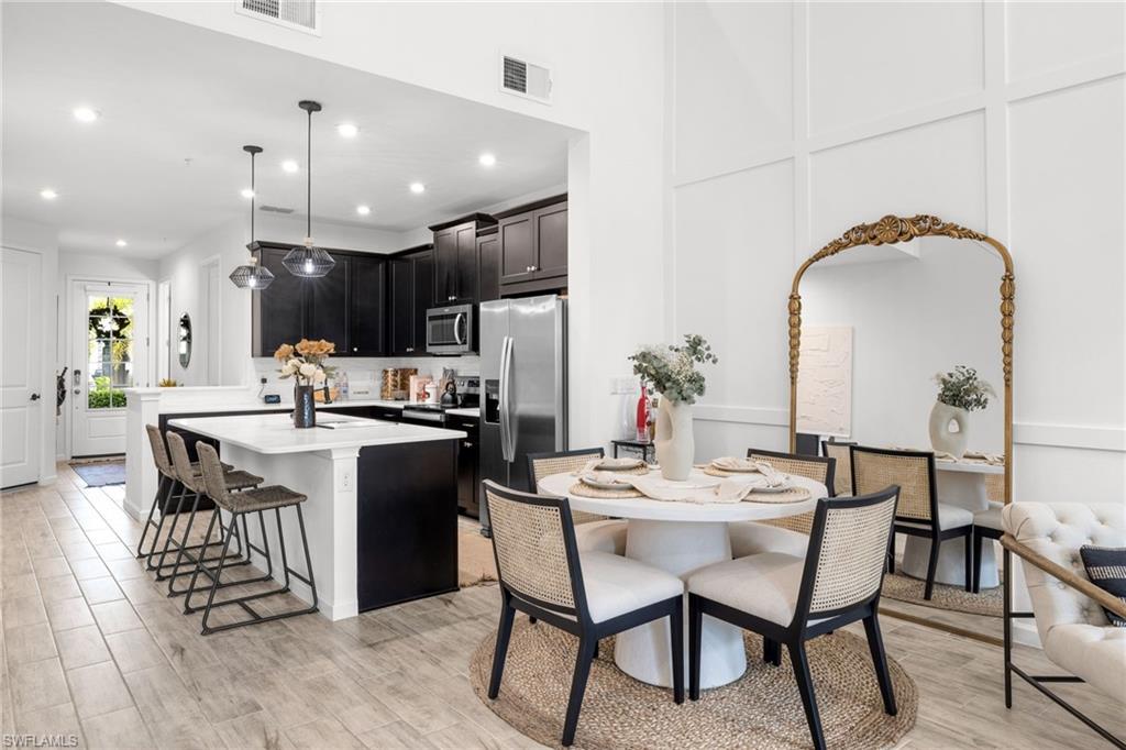 Open-concept living space featuring wood-finish flooring, a kitchen with black cabinetry and stainless steel appliances, a kitchen island with seating, a dining area, and a white paneled wall