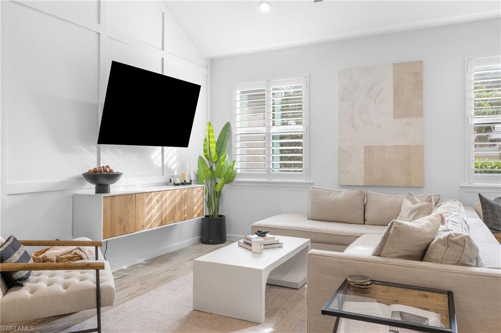 Living area featuring vaulted ceilings with accent paneling, wood-finish flooring, two large windows with plantation shutters, and a floating entertainment console with wood-panel doors