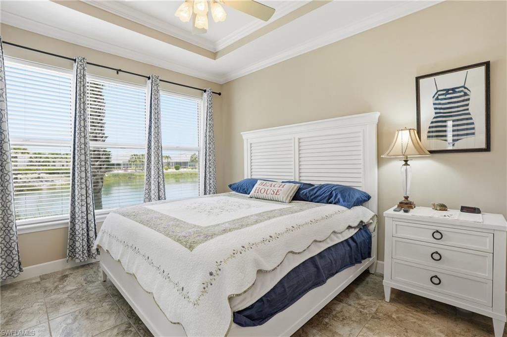 Spacious bedroom featuring three large windows with water scenery, tray ceiling, ceiling fan with light fixture, and tile flooring