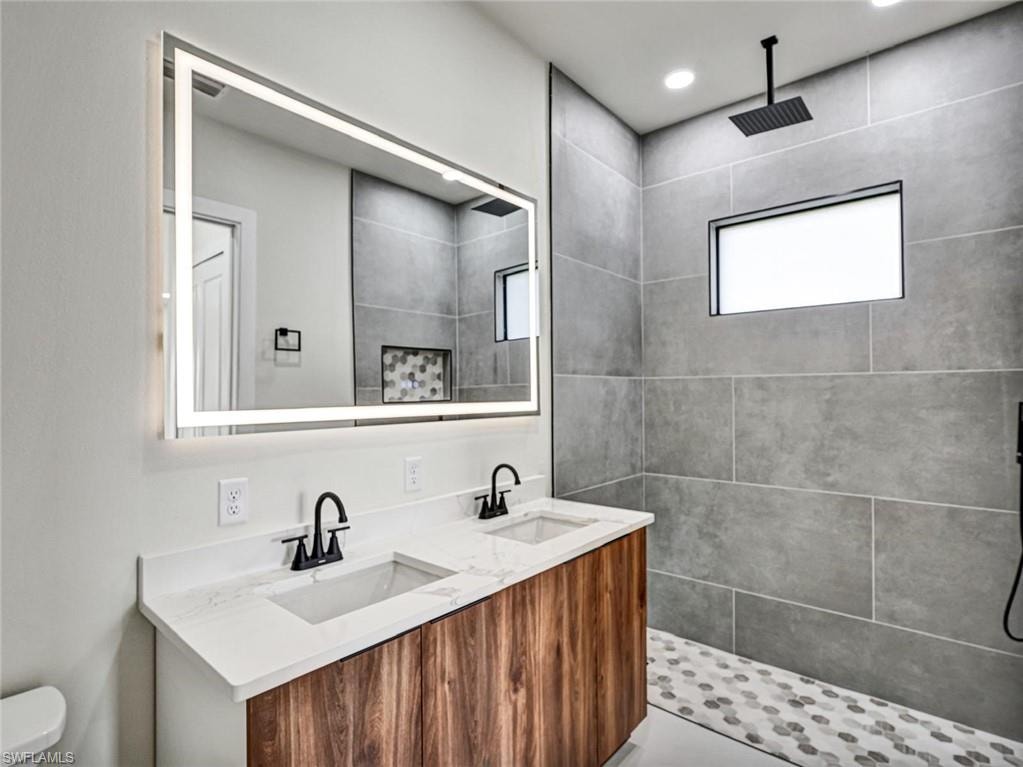 Bathroom featuring a dual-sink vanity with a wood-finish cabinet and white countertop, illuminated mirror, and a walk-in shower with large format tile walls, a rainfall showerhead, and a mosaic tile floor