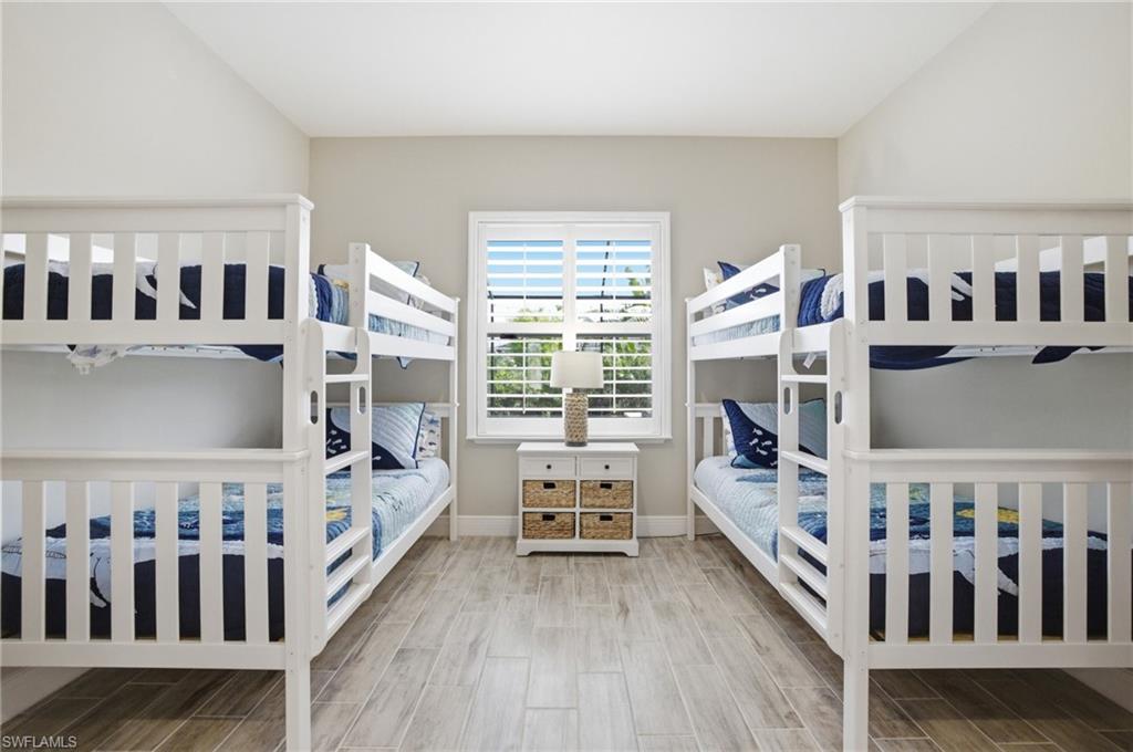 Spacious room featuring two sets of white bunk beds, light wood-finish flooring, and a central window with white plantation shutters