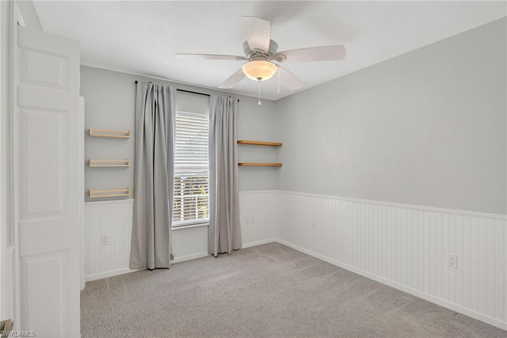 Carpeted interior room featuring a window with blinds and drapes, ceiling fan with light fixture, and wainscoting with wall shelving