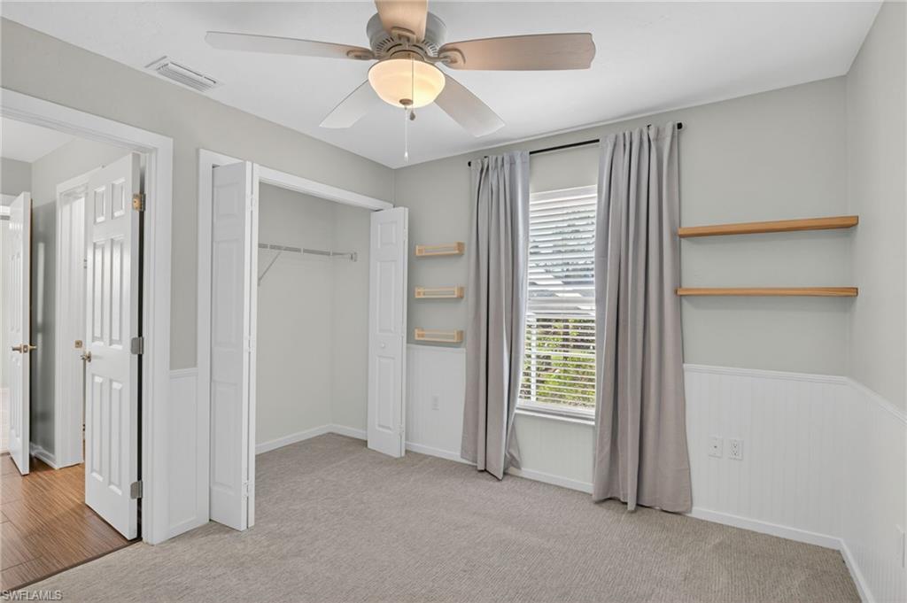 Carpeted room featuring a ceiling fan with light fixture, window with blinds and drapes, board and batten wall detailing, and built-in floating shelves