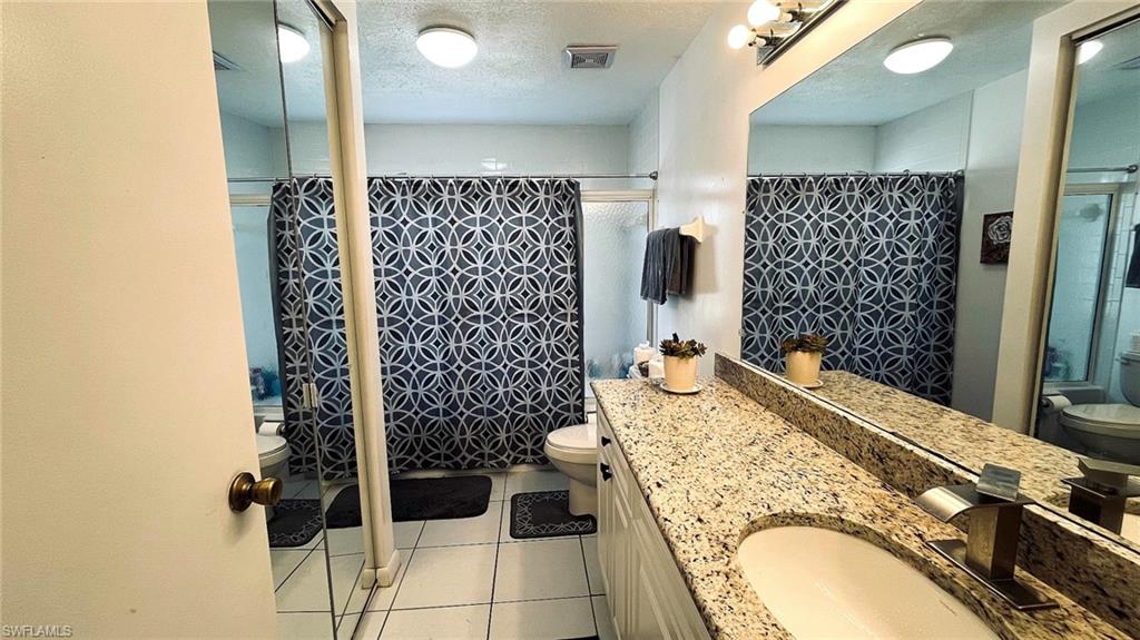 Bathroom featuring a granite-finish vanity with an undermount sink, a large wall-mounted mirror, white tile flooring, and a shower/tub combination behind a patterned curtain