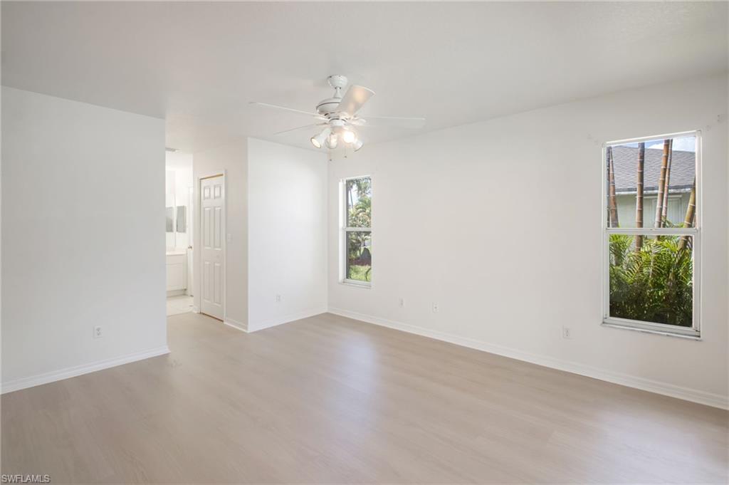 Spacious room featuring light wood-finish flooring, a white ceiling fan with integrated lighting, multiple windows providing natural illumination, and crisp white walls