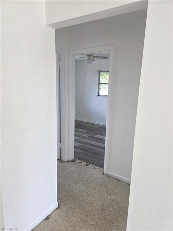 Interior hallway with light-colored carpeting, leading to a room featuring wood-finish flooring, a window, and a ceiling fan