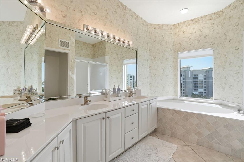 Spacious bathroom featuring a built-in soaking tub with tiled surround, dual vanity with white cabinetry, large wall-mounted mirror, and patterned wallpaper