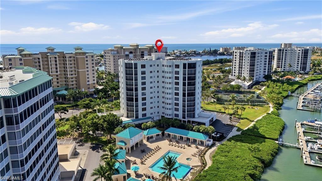 High-rise condominium with multiple balconies, a ground-level pool deck with two swimming pools, surrounded by mature landscaping and a marina with boat slips
