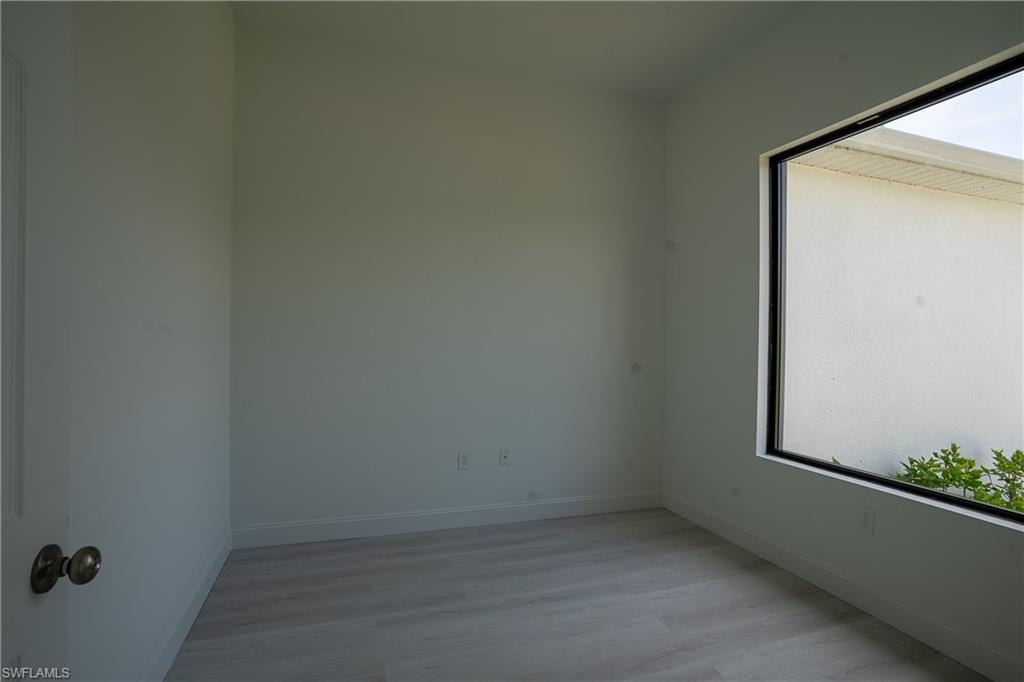 Bright room featuring a large black-framed window, light wood-finish flooring, white baseboards, and a white interior door with brass hardware