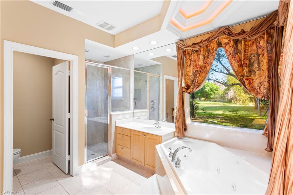 Spacious bathroom featuring a soaking tub, separate glass-enclosed shower, dual vanity with light wood cabinetry, recessed lighting, and a large window overlooking a landscaped expanse