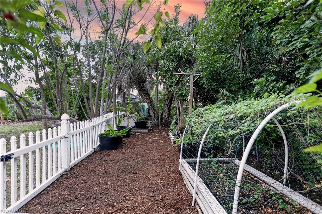 Garden path with a white picket fence, raised garden beds with trellises, and mature trees