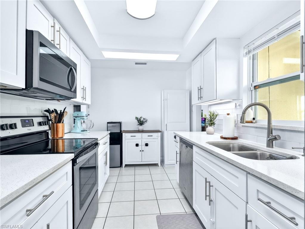 Galley kitchen featuring white shaker-style cabinetry, light-toned countertops, a white subway tile backsplash, and a double basin stainless steel sink with a gooseneck faucet