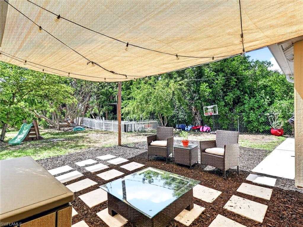 Outdoor patio featuring a light-colored shade sail with string lighting, a paver patio with wood chip accents, and a large grassy yard enclosed by a white picket fence and chain-link fencing