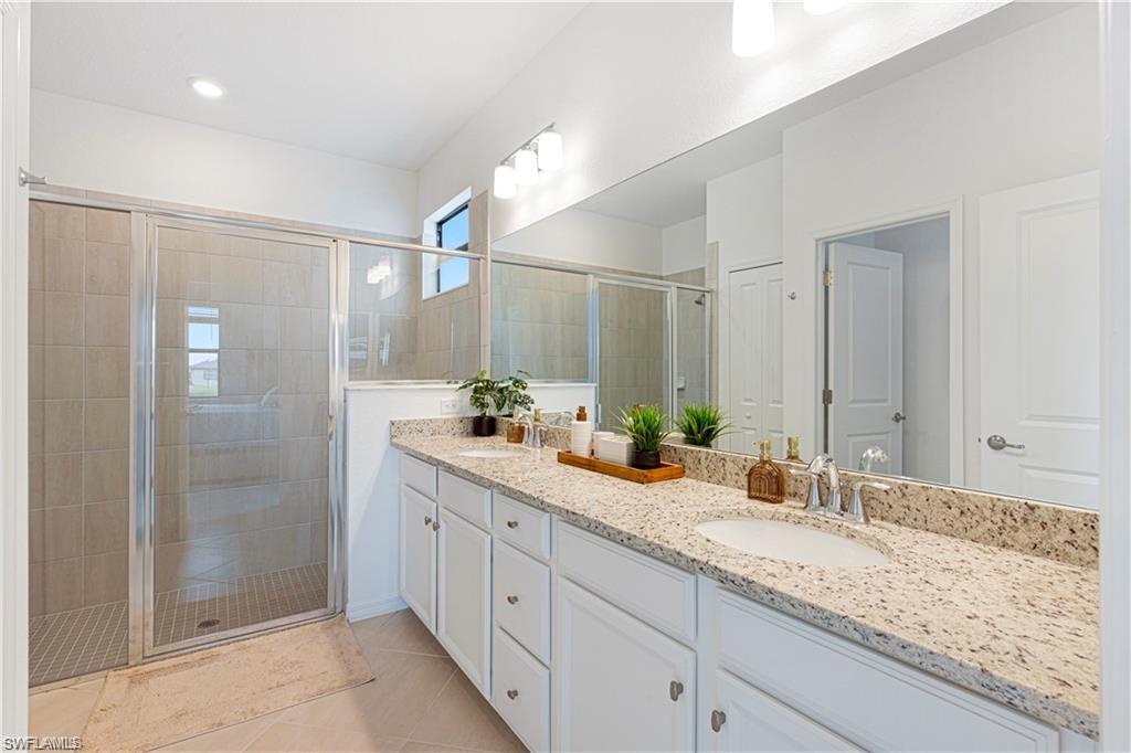 Bathroom featuring a double vanity with light-toned countertops, dual undermount sinks with polished chrome fixtures, white cabinetry, a large frameless mirror, and an enclosed shower with full height tiling and a glass door