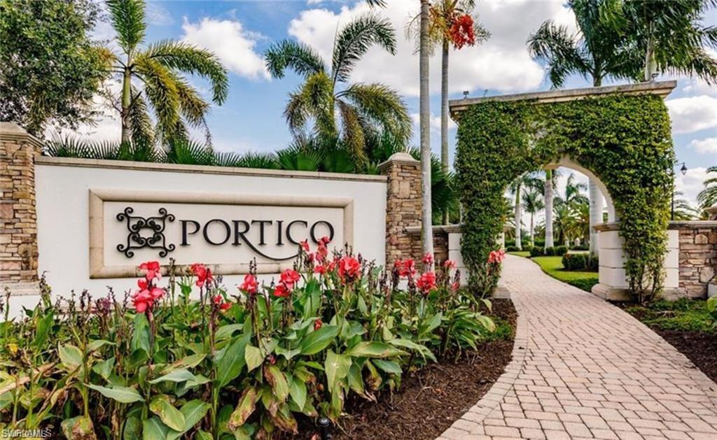 Community entrance featuring a stone-clad sign with decorative ironwork, a brick paver pathway, a vine-covered archway, and lush tropical landscaping with palm trees and vibrant red flora