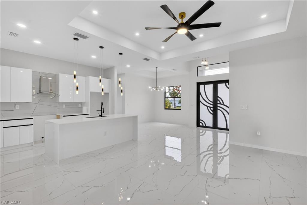 Open-concept living area featuring large format white tile flooring with veining, a contemporary kitchen island with pendant lighting, white high-gloss cabinetry, and a black ceiling fan with gold accents