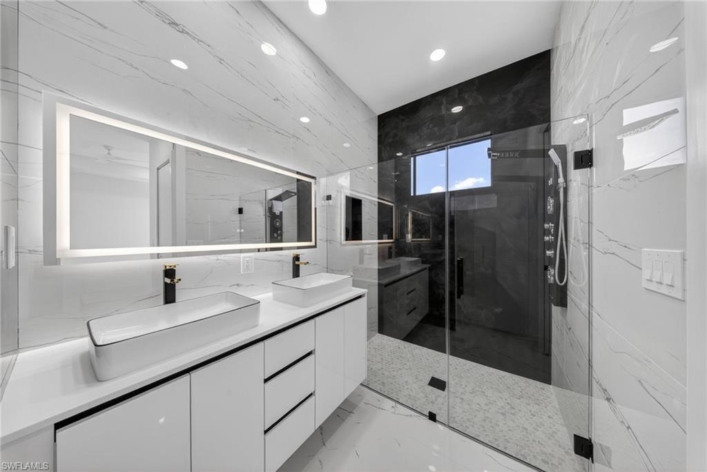 Contemporary bathroom featuring a double vanity with vessel sinks, an illuminated mirror, a spacious glass-enclosed shower, dark large-format tile accents, and integrated overhead lighting