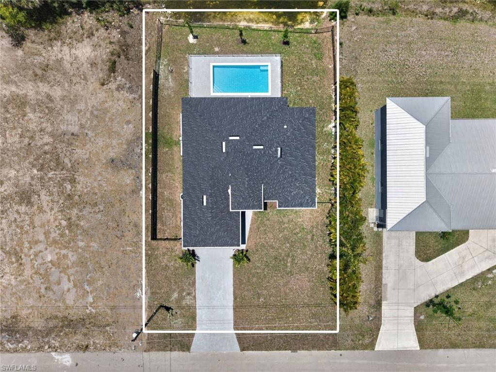 Aerial view showcasing the property's rectangular lot with a rectangular swimming pool, a dark shingle roof, a light-colored driveway, and surrounding natural landscaping