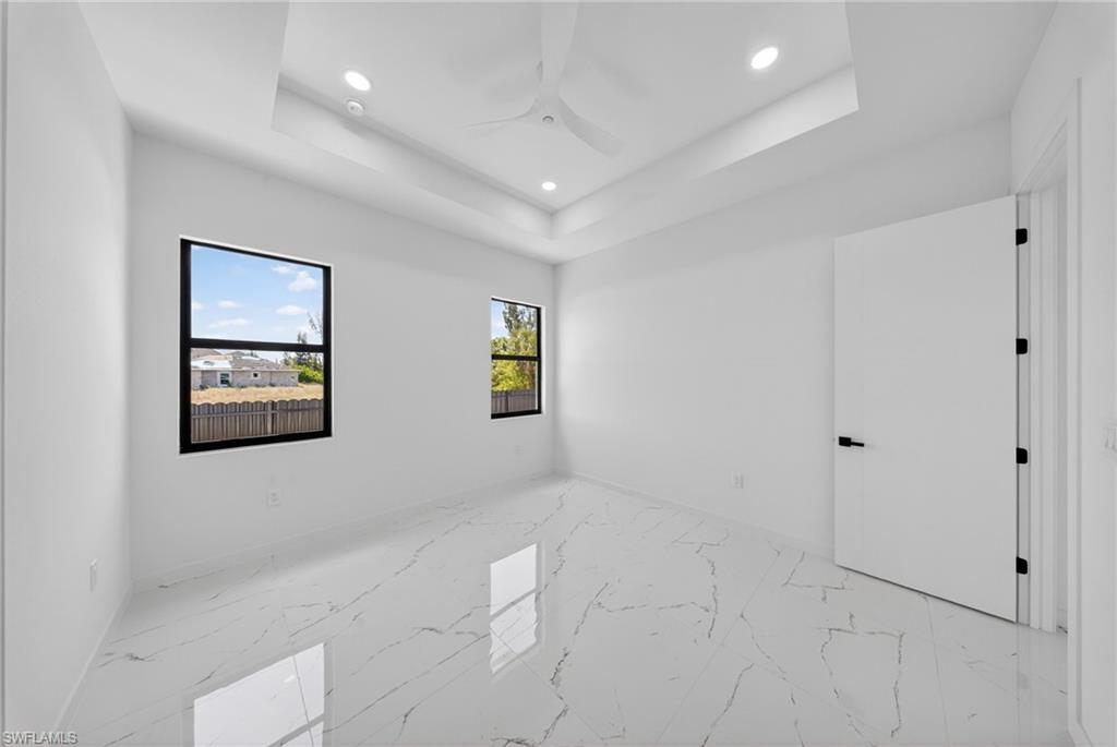 Spacious interior room featuring a tray ceiling with recessed lighting and a ceiling fan, white marble-look tile flooring, and two black-framed windows