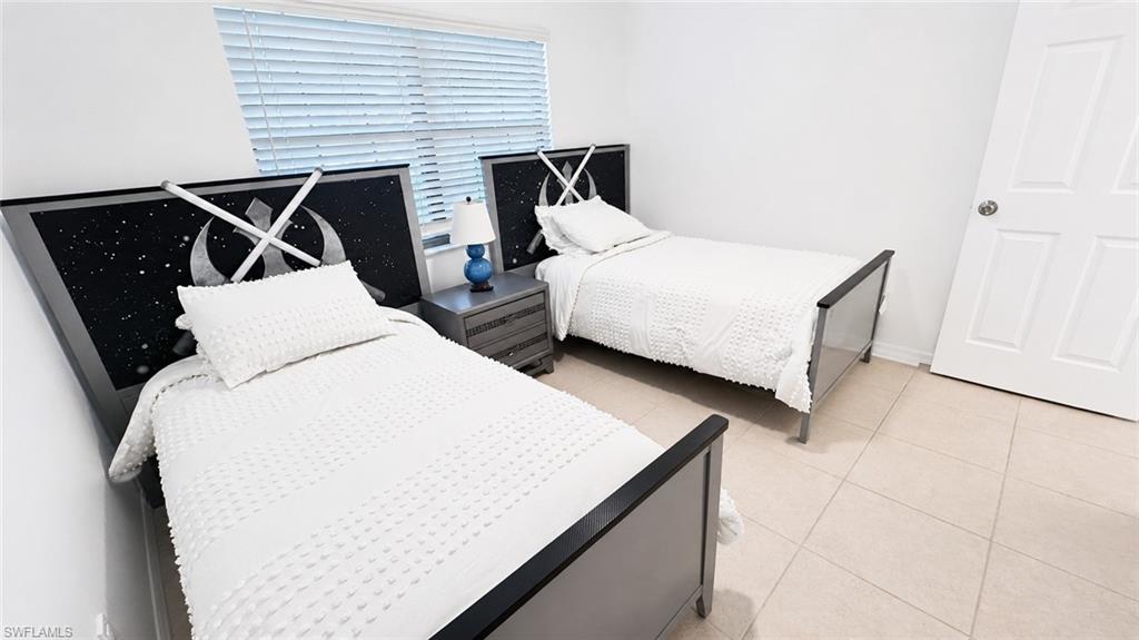 Room featuring two beds with themed headboards, neutral tile flooring, a window with blinds, a side table, and a white paneled door
