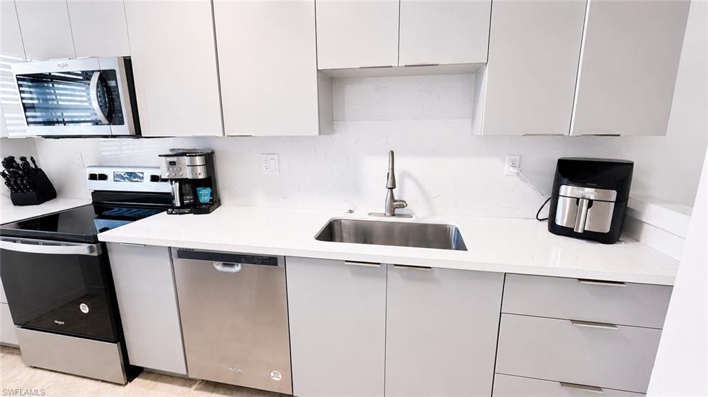 Modern kitchen featuring light gray flat-panel cabinetry, white countertops, stainless steel appliances, a stainless steel sink with a gooseneck faucet, and a white backsplash