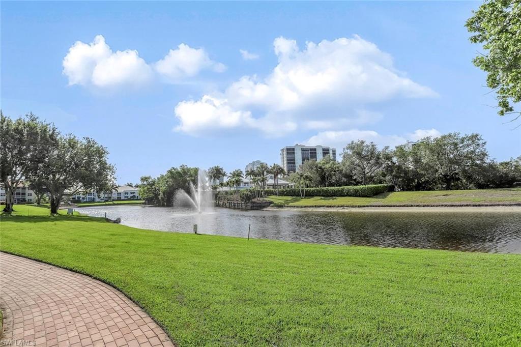 Expansive waterfront setting featuring a tranquil fountain, lush green lawn, mature trees, and a paver pathway