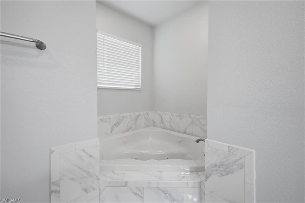 Corner jetted tub featuring a tile surround with marble-look accents, a window with horizontal blinds, and a wall-mounted towel bar