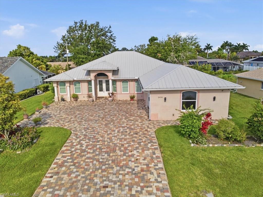 Exterior residence featuring a multicolored paver driveway, light-colored stucco facade, arched entry with double doors, and a metal roof