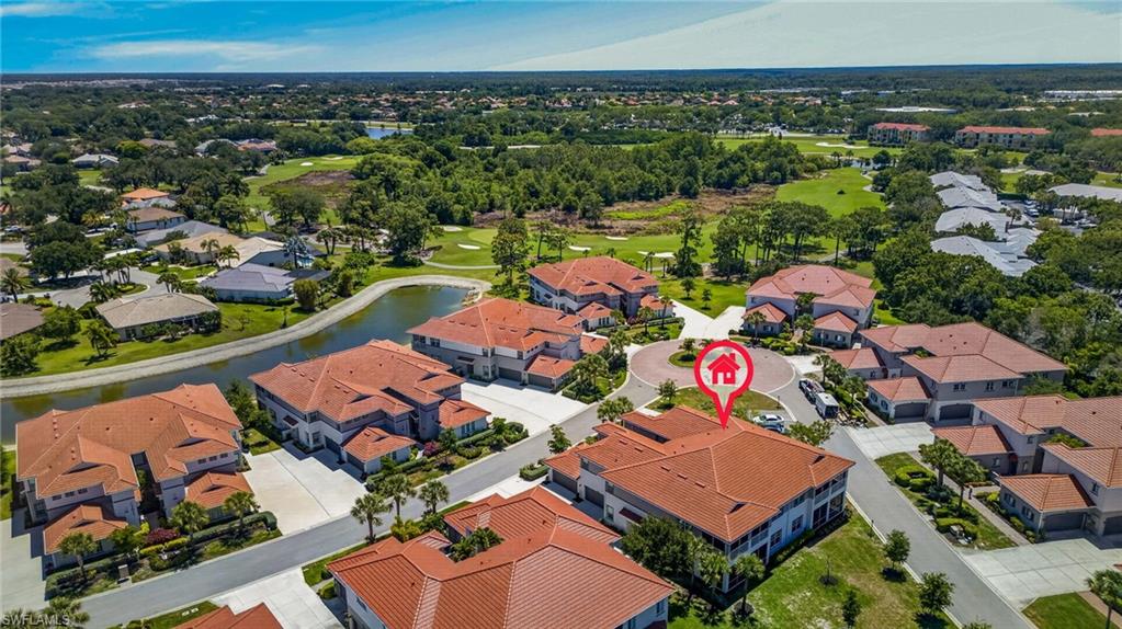 Aerial perspective of a residential community featuring red tile roofs, multiple buildings with balconies, mature landscaping including palm trees, and water features