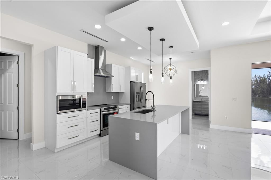Kitchen with hanging light fixtures, stainless steel appliances, a center island with sink, white cabinets, and light stone counters