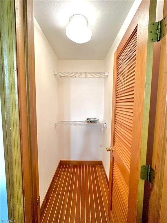 Interior closet featuring wood-finish flooring, two wire shelves, a flush-mount ceiling light, and a louvered wood door with brass hardware