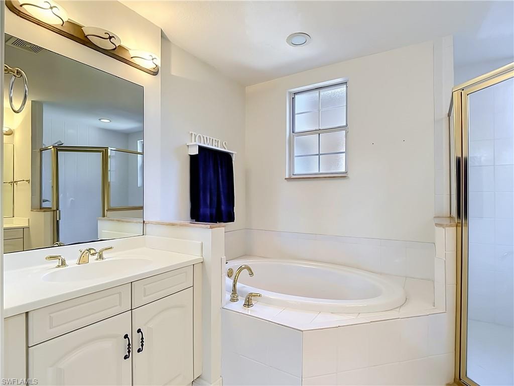 Bathroom featuring a white built-in soaking tub with gold-finish fixtures, a dual vanity with white cabinetry and integrated sinks, and a separate glass-enclosed shower with gold-finish trim
