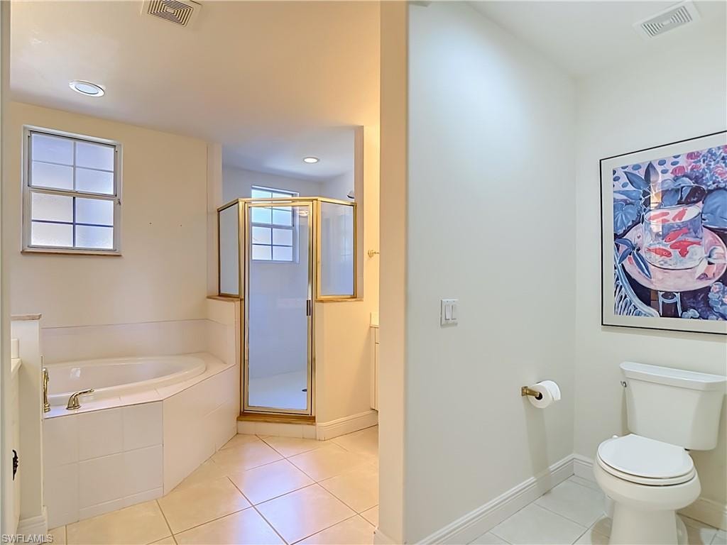 Bathroom featuring a corner soaking tub with deck-mounted faucet, a separate glass-enclosed shower with gold-tone framing, a commode, and white tile flooring