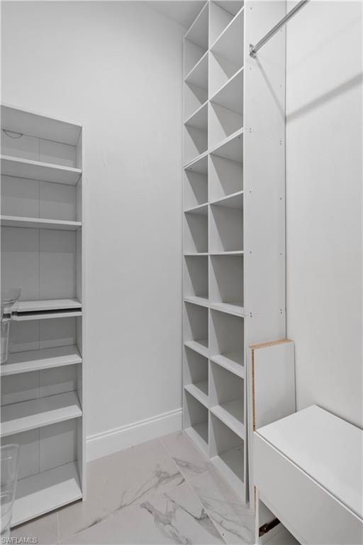 Walk-in closet featuring white built-in shelving, white baseboards, a chrome hanging rod, and light-toned tile flooring with subtle veining