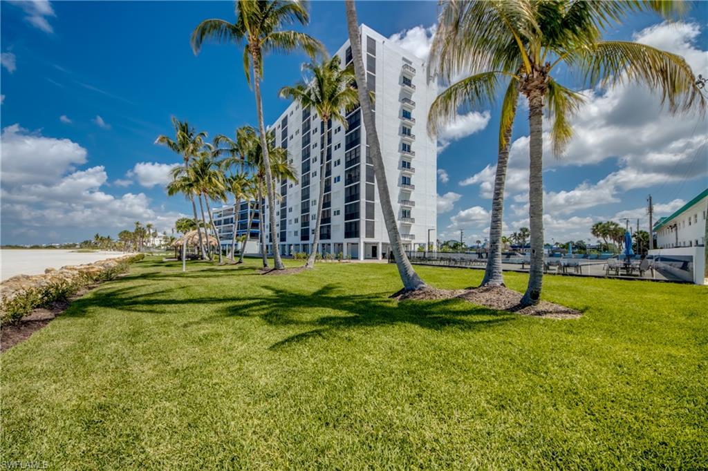 Waterfront property featuring a multi-story building, lush green lawn, mature palm trees, sandy beach access, and a boat dock