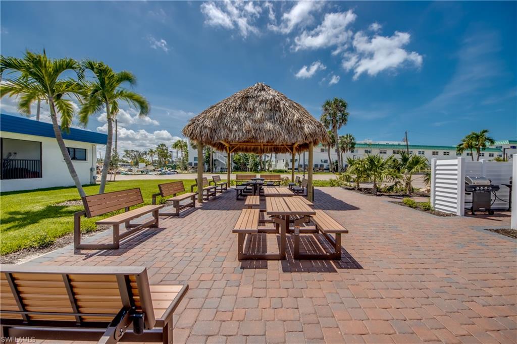 Expansive paved patio area featuring a large thatched-roof gazebo, multiple picnic tables, and additional seating benches