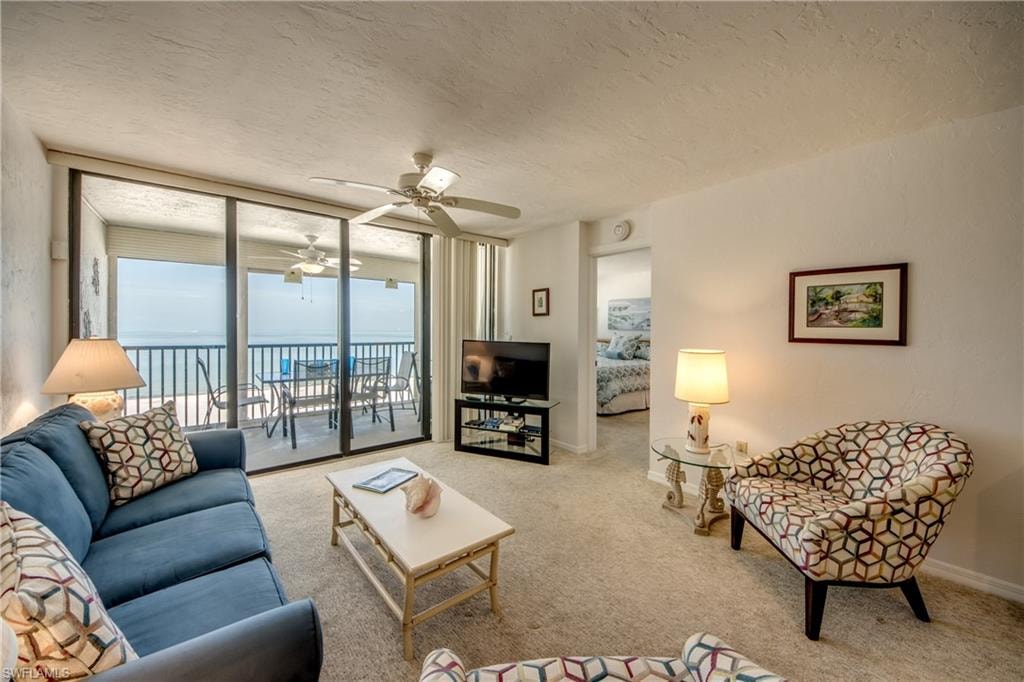 Living area with sliding glass doors opening to a balcony, featuring a ceiling fan, textured white walls, and neutral carpet flooring