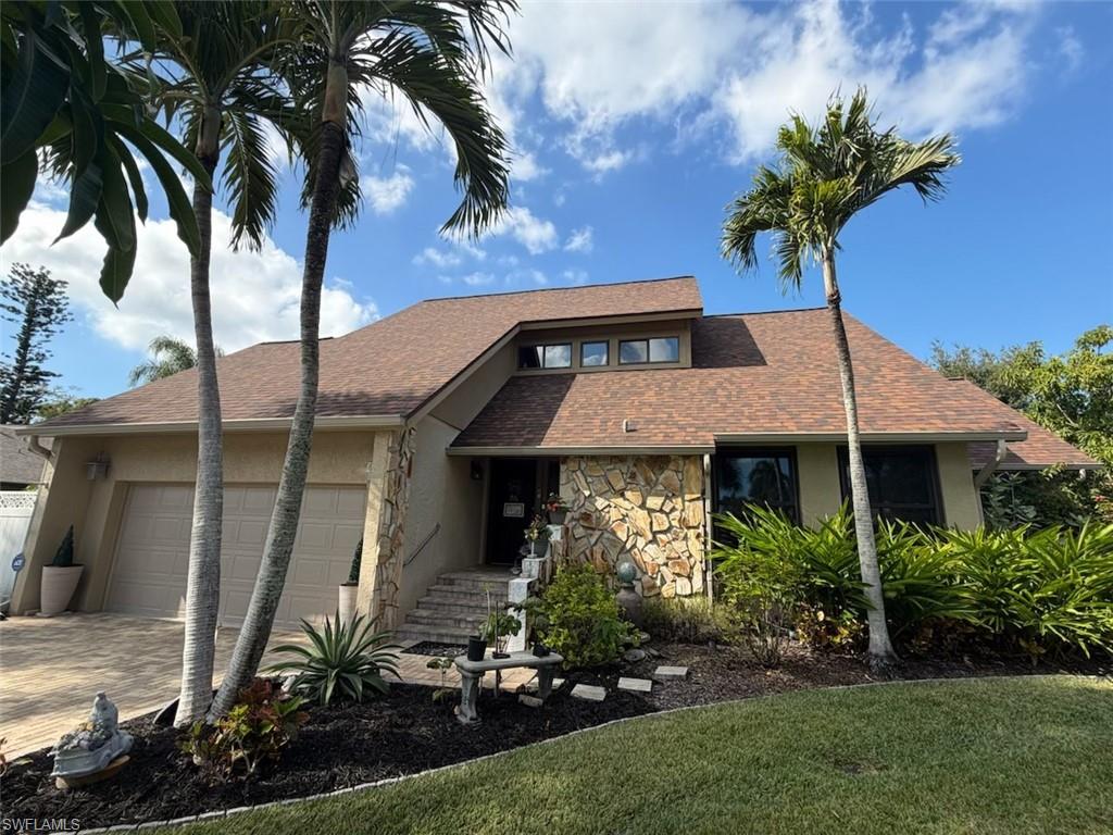 Stucco exterior with stone accent facade, featuring a multi-level roofline, integrated two-car garage, mature tropical landscaping, and a paved driveway