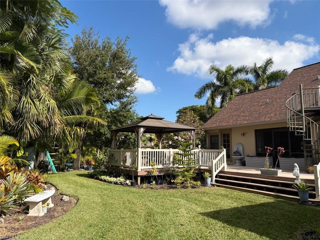Expansive backyard featuring a manicured lawn, a raised wooden deck with a gazebo, mature palm trees, a spiral staircase, and a stone bench