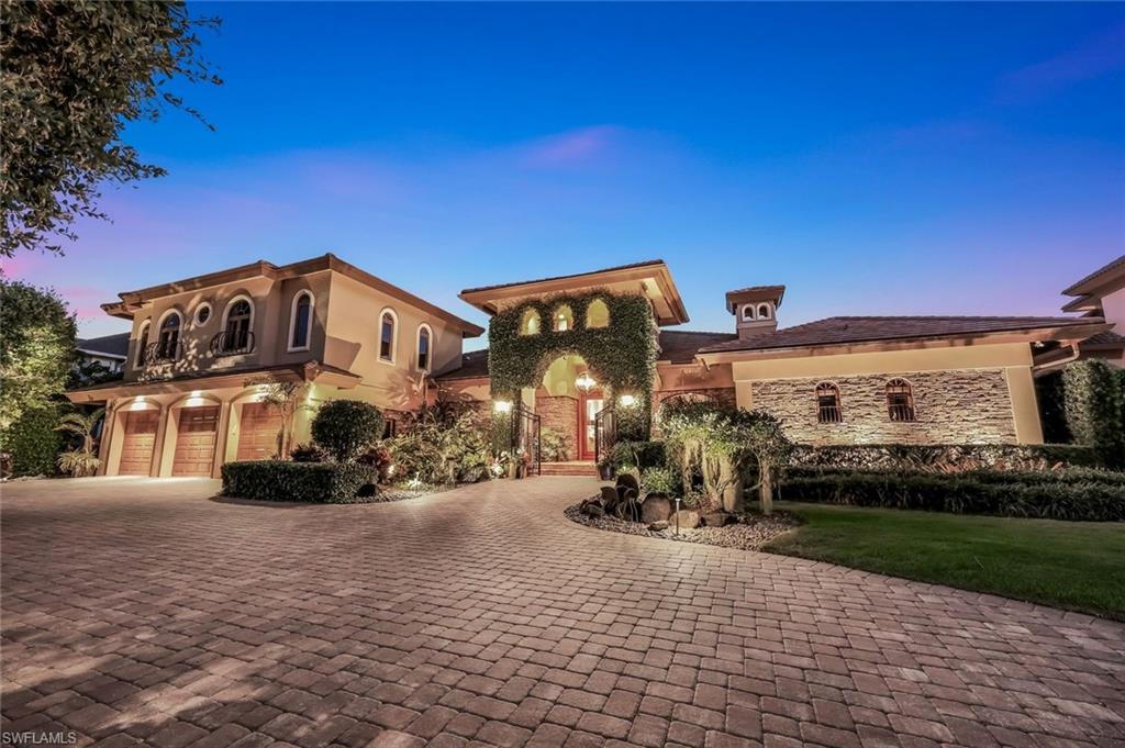 Mediterranean-inspired architecture featuring a paver driveway, three-car garage with wood doors, stone facade accents, and lush landscaping with up-lighting