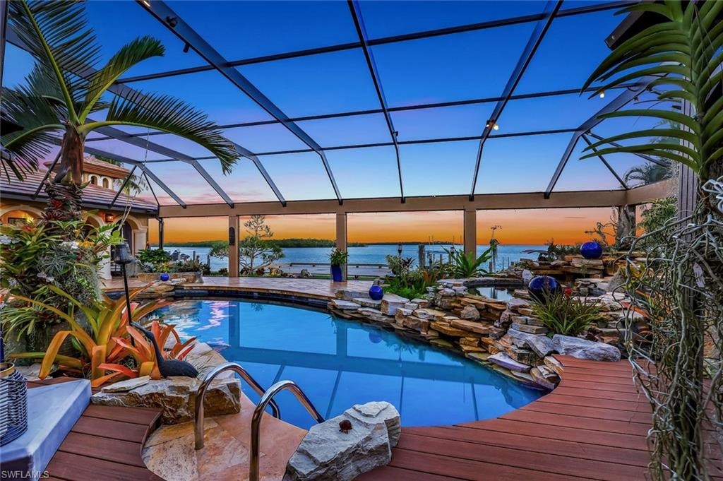 Screened pool enclosure featuring a lagoon-style pool, rock water features, wood-finish decking, and tropical landscaping