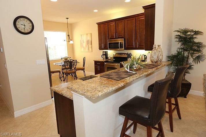 Kitchen with a kitchen bar, appliances with stainless steel finishes, light stone counters, hanging light fixtures, and light tile patterned floors