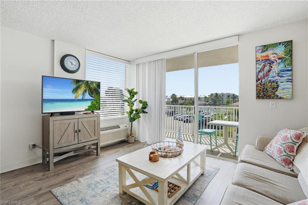 Living area featuring wood-finish flooring, a wall-mounted clock, a window with horizontal blinds, and sliding glass doors opening to a balcony - Virtually Edited Image