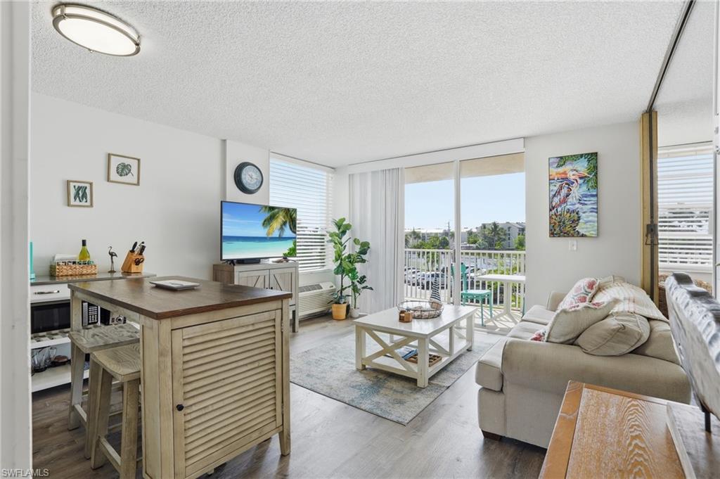 Open-concept living space featuring wood-finish flooring, a sliding glass door to an exterior balcony, and a built-in kitchen island with seating - Virtually Edited Image