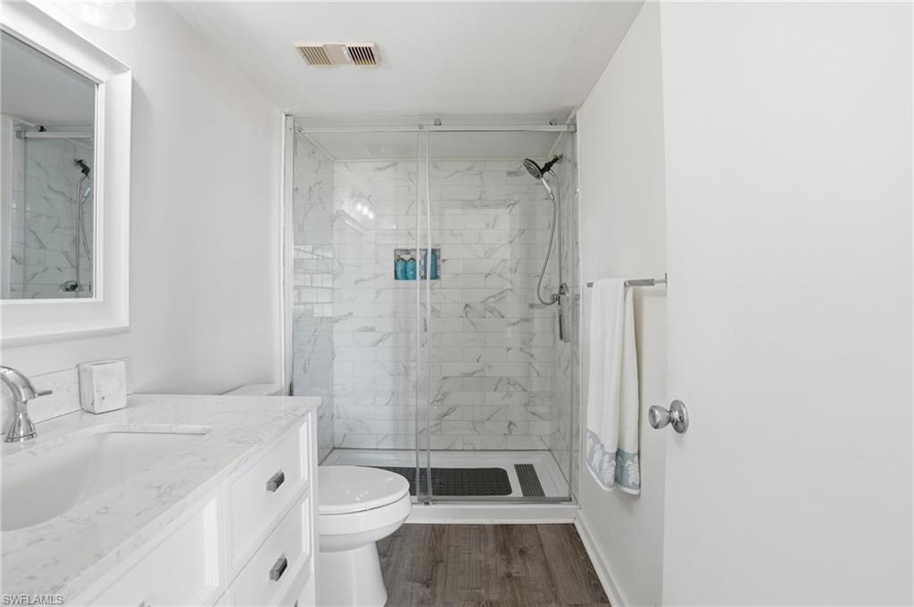 Bathroom featuring a glass-enclosed shower with subway tile surround, a single vanity with an undermount sink and light-toned countertop, wood-finish flooring, and a framed mirror - Virtually Edited Image