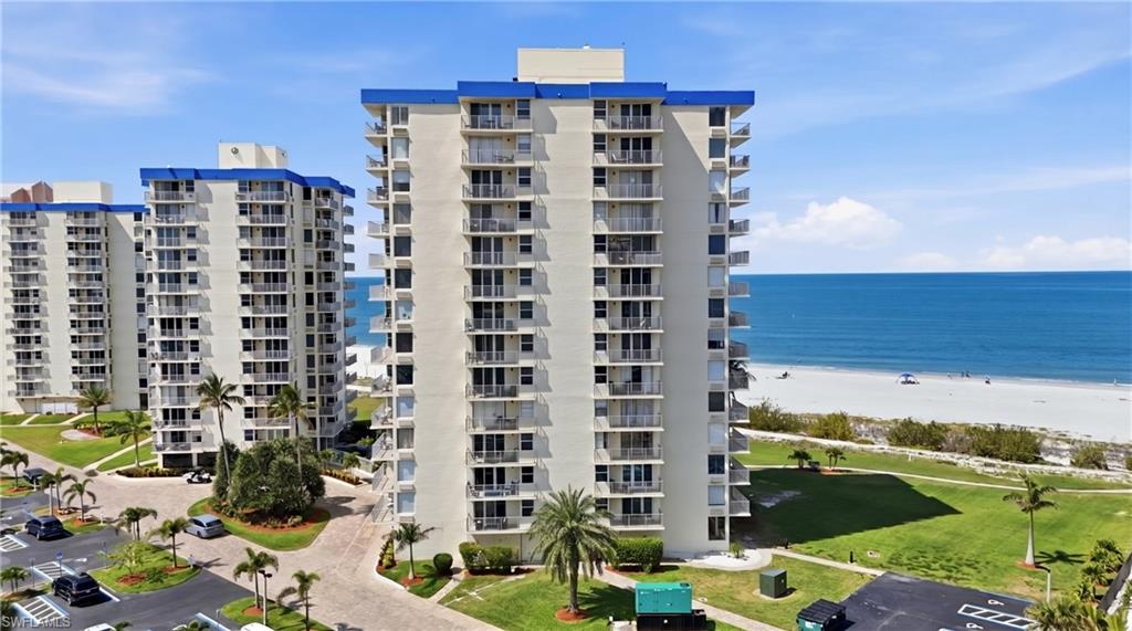 High-rise condominium building featuring multiple levels of private balconies, direct beach access, coastal water frontage, and surrounding green spaces with mature palm trees - Virtually Edited Image